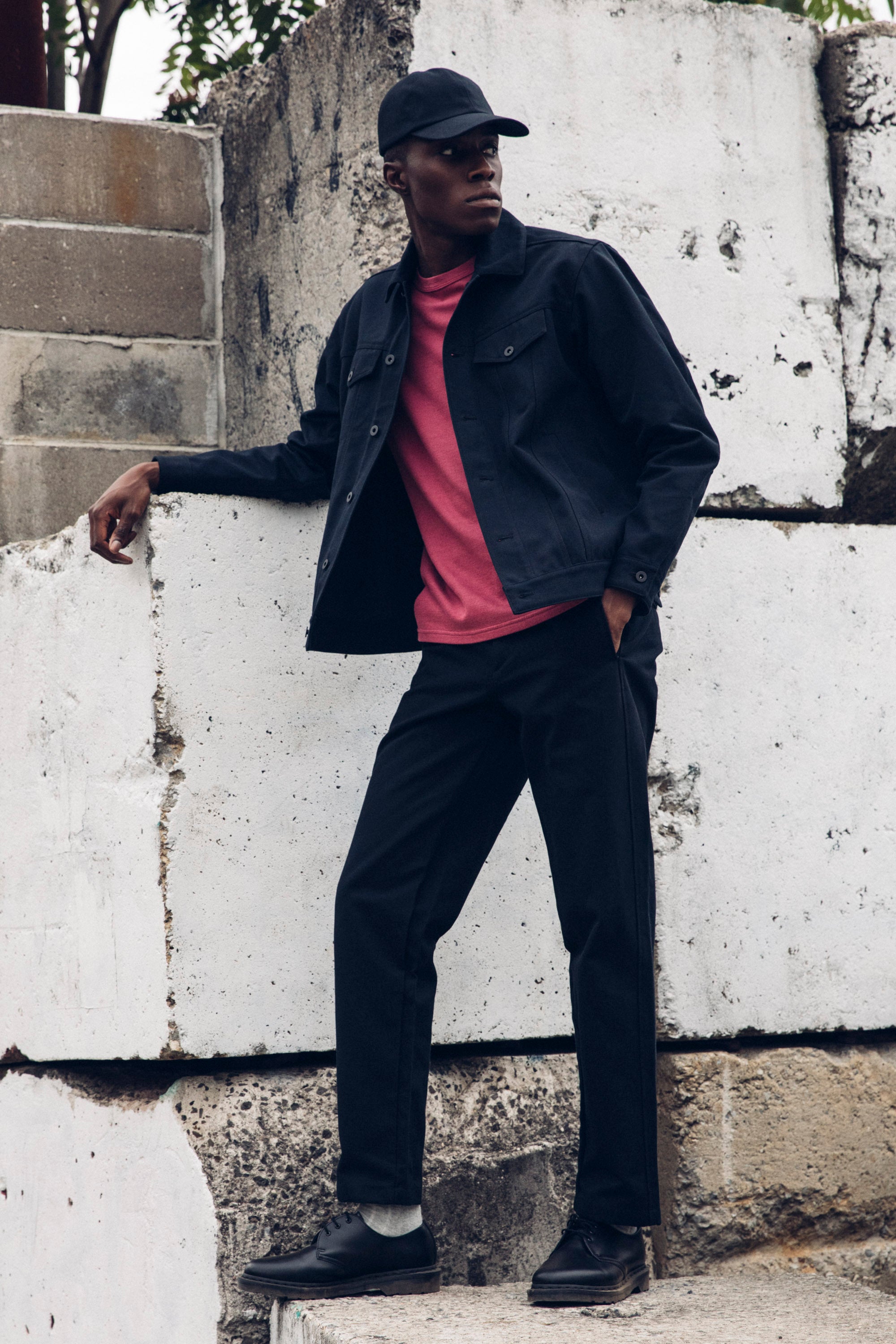 Edem in the Washed Black Duckworks with a Duckcap and a Duckcloth Shank Jacket, outdoors leaning on concrete blocks