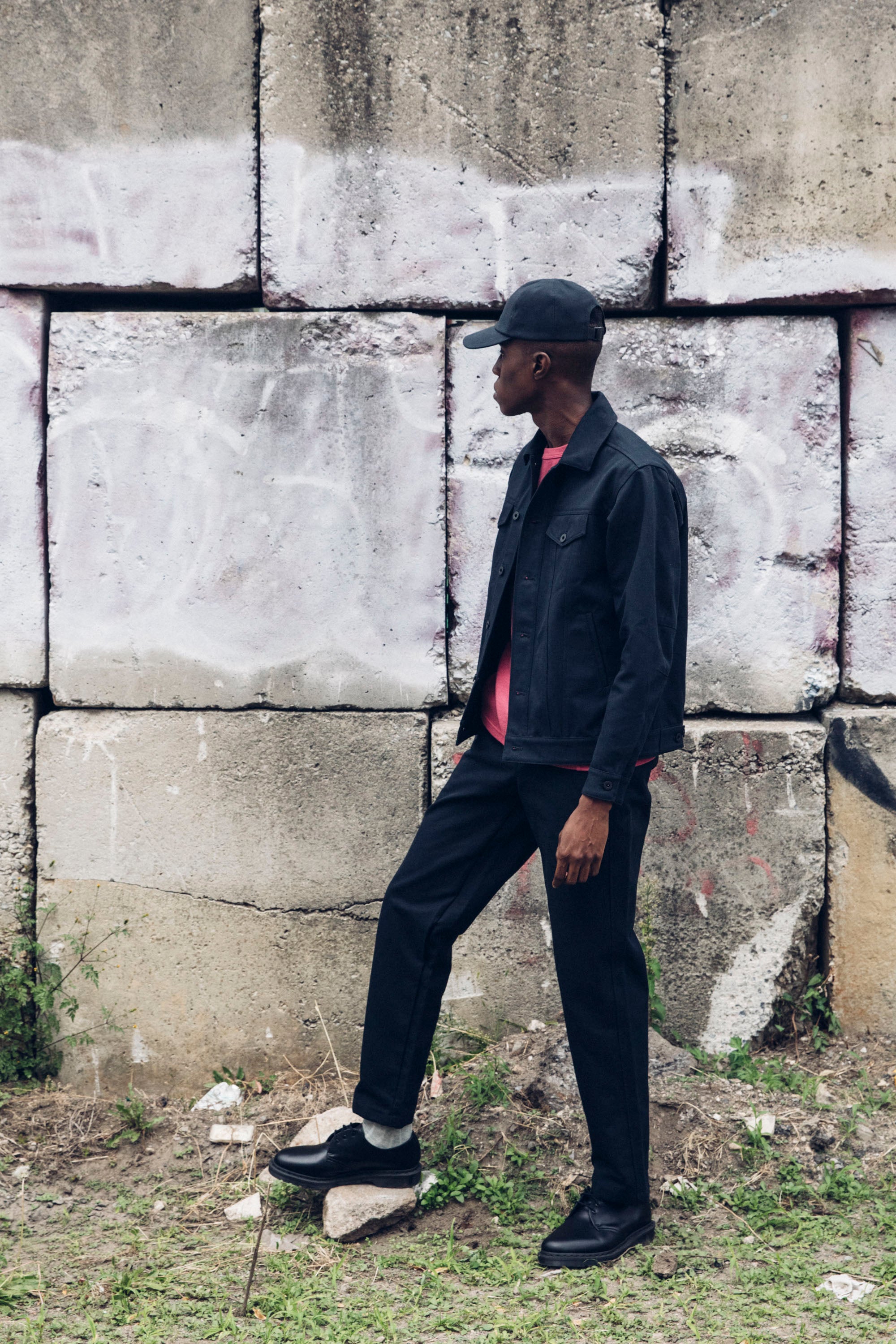 Edem in the Washed Black Duckworks with a Duckcap and a Duckcloth Shank Jacket, outdoors in front of concrete blocks